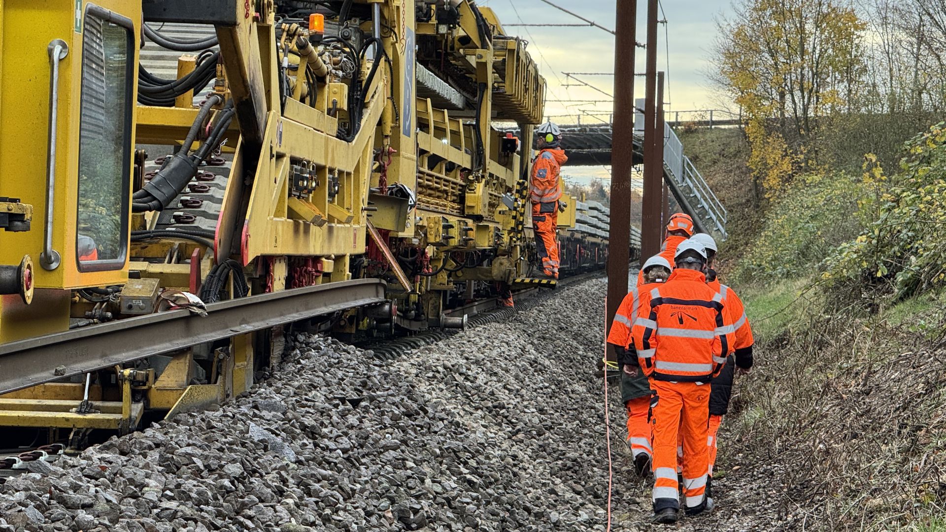 Medarbejdere går langs sporet, mens sporombygningsmaskinen løfter jernbaneskinnerne og udskifter den gamle sveller.